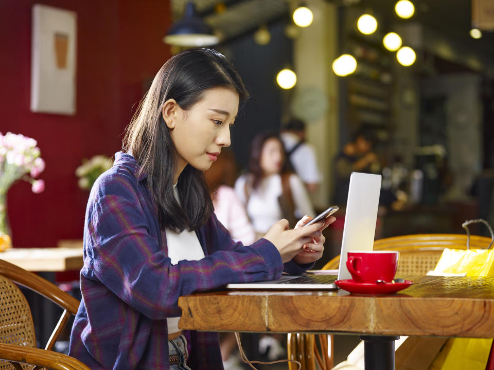 A young woman checks her phone and laptop in a coffee shop.