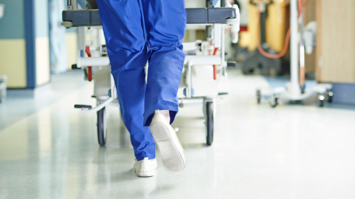  Close up on legs of medic wearing scrubs and walking with a gurney along hospital corridor. 