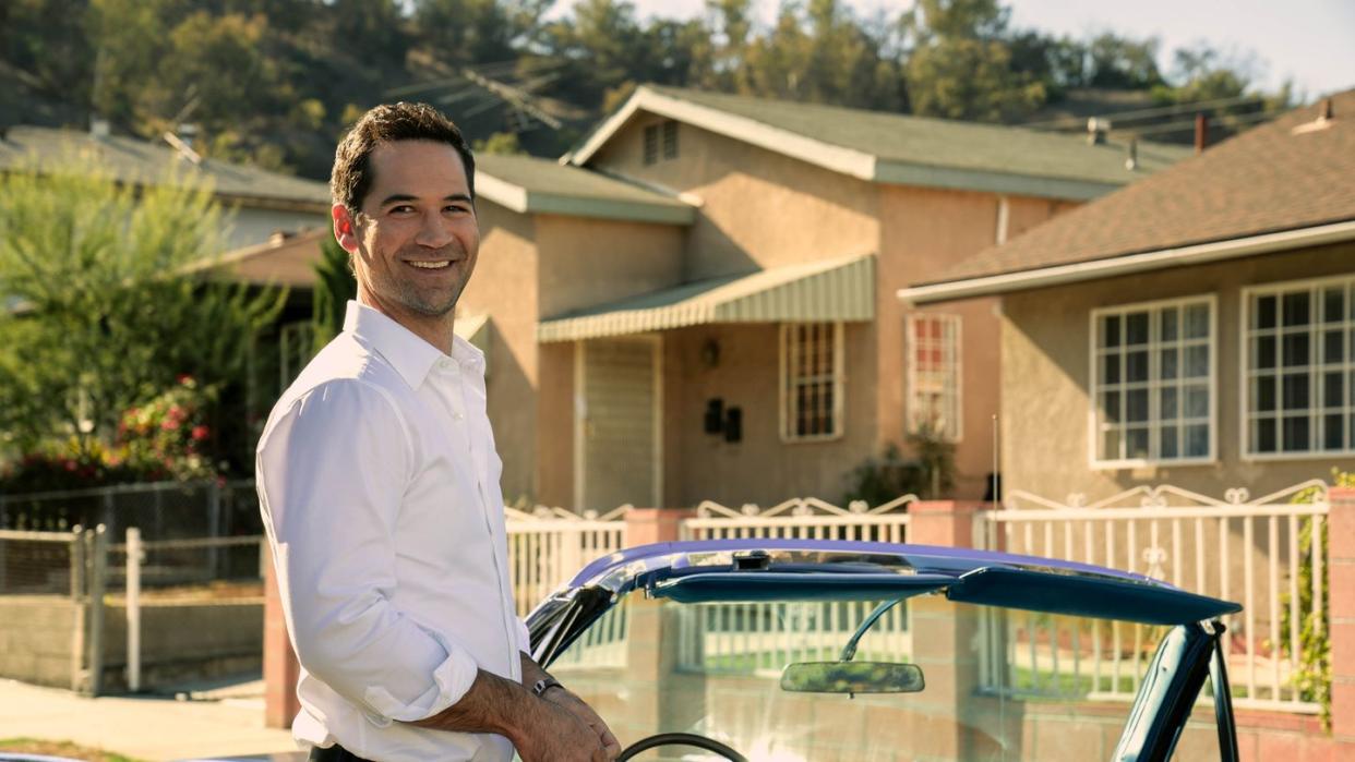 manuel garcia rulfo as mickey haller in lincoln lawyer season 2 smiling as he stands beside vintage blue lincoln convertible