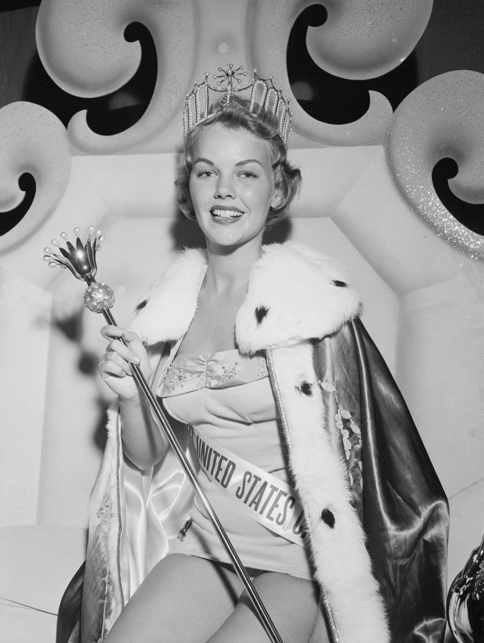 Miss USA 1955 Carlene King Johnson sits on a throne with a tiara, scepter, and robe.