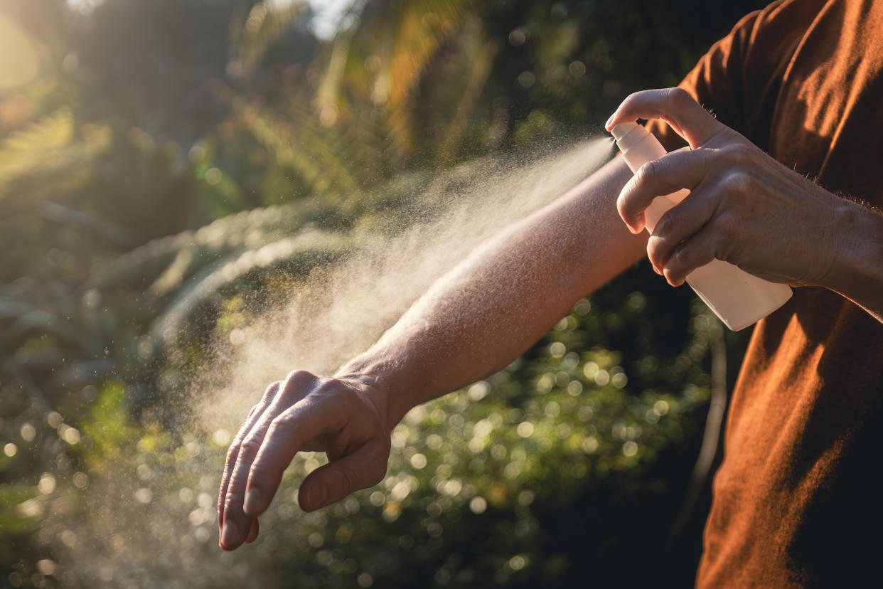 A man in a T-shirt sprays bug spray onto his forearm outdoors.