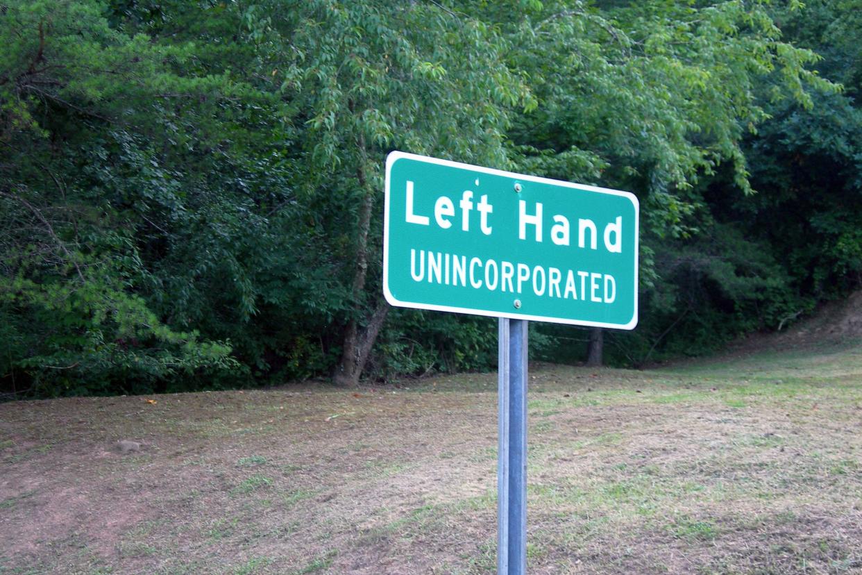 Green road sign 'Left Hand, Unincorporated' along the side of the road, welcoming those entering Left Hand, West Virginia, with grass and forest in the background