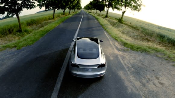 A silver Model 3 driving on an open road with trees lining it on each side