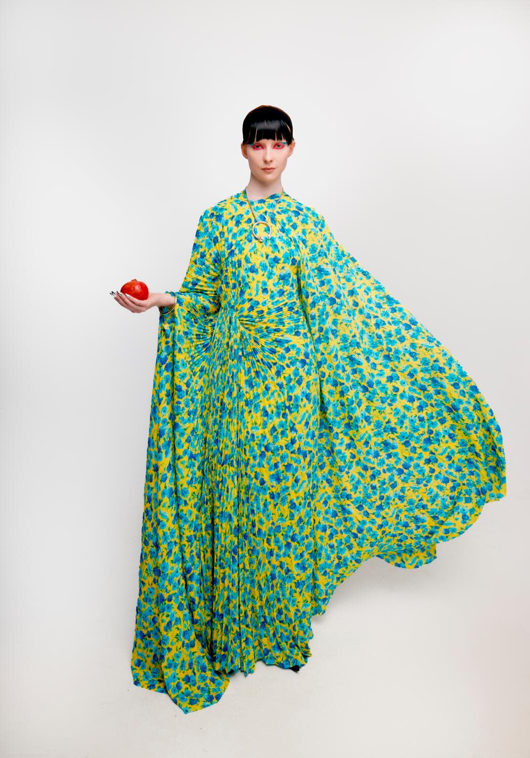 A woman wears a flowery dress and holds a pomegranate.