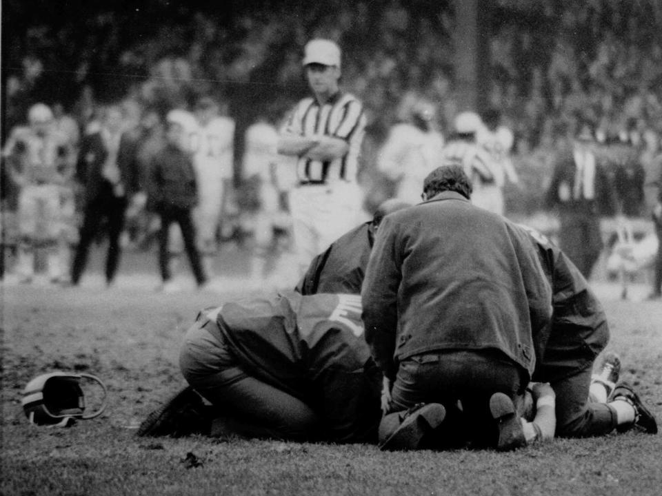 a black and white image of a people kneeling around a football player on a field, depicting the 1971 fatal collapse of Chuck Hughes during an NFL game.