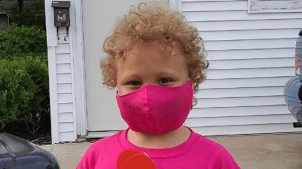 Jurnee Hoffmeyer, wearing a pink face mask, stands near a home with a mailbox and door visible.