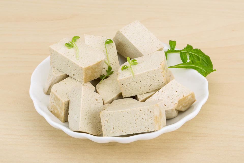 A white plate holds several cubes of tofu garnished with small green leaves, presented on a light wooden surface