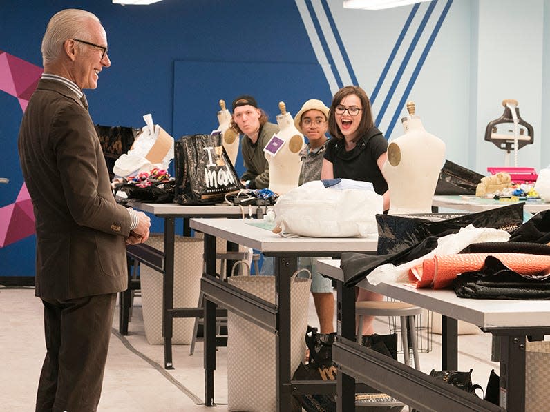 The writer sits at her work table and laughs while Tim Gunn stands at front of room