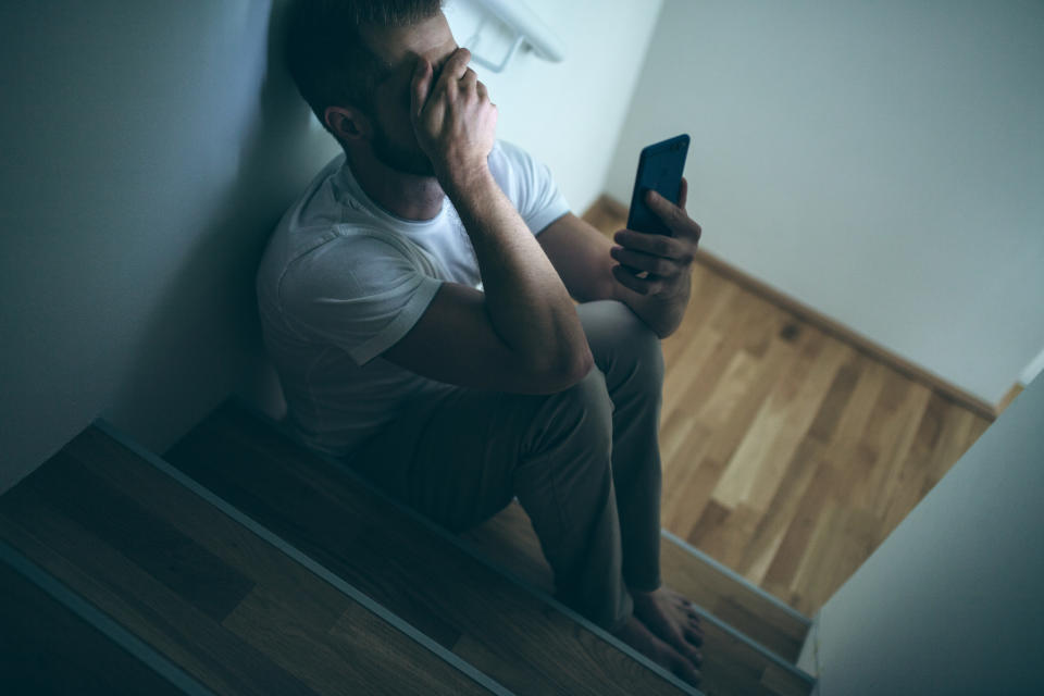 Depressed man sitting in the dark with his smartphone to represent the cost of living putting increased pressure on him.
