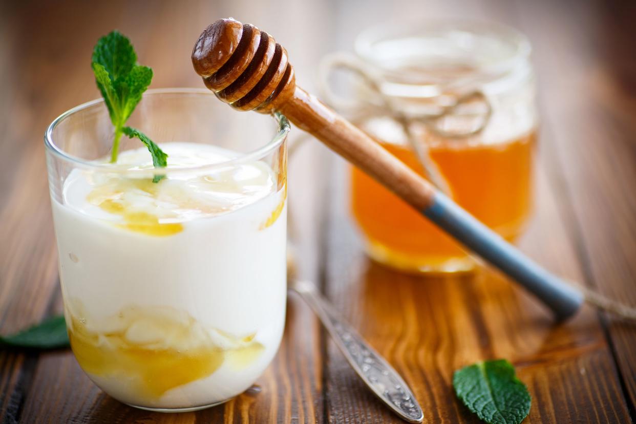 Greek yogurt with honey and cinnamon in a glass dessert cup, with a honey dipper leaning on it, next to a spoon, on a wooden table with a glass jar of honey in the background