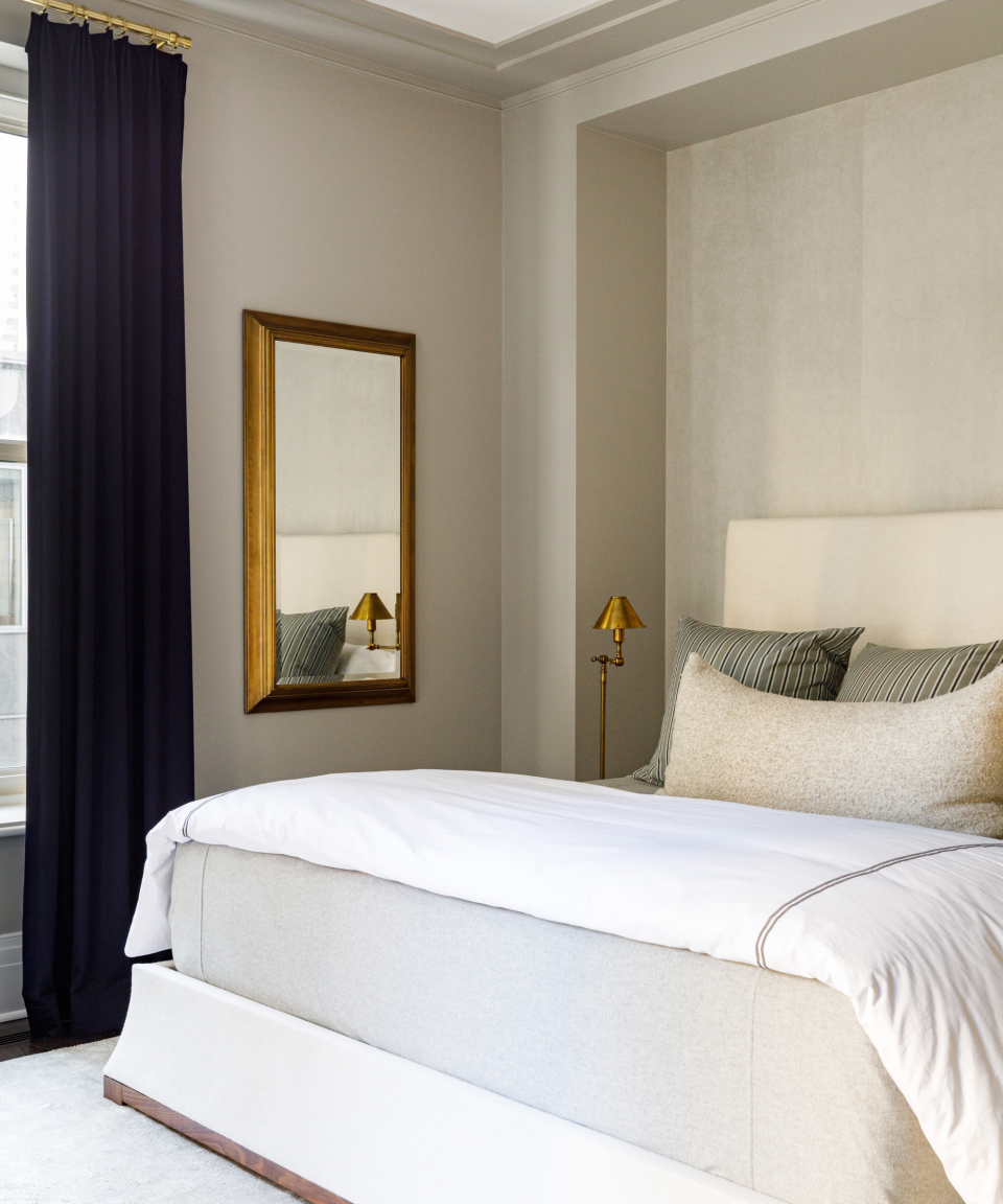 Neutral bedroom with brass-framed wall mirror, throw pillows and white sheets