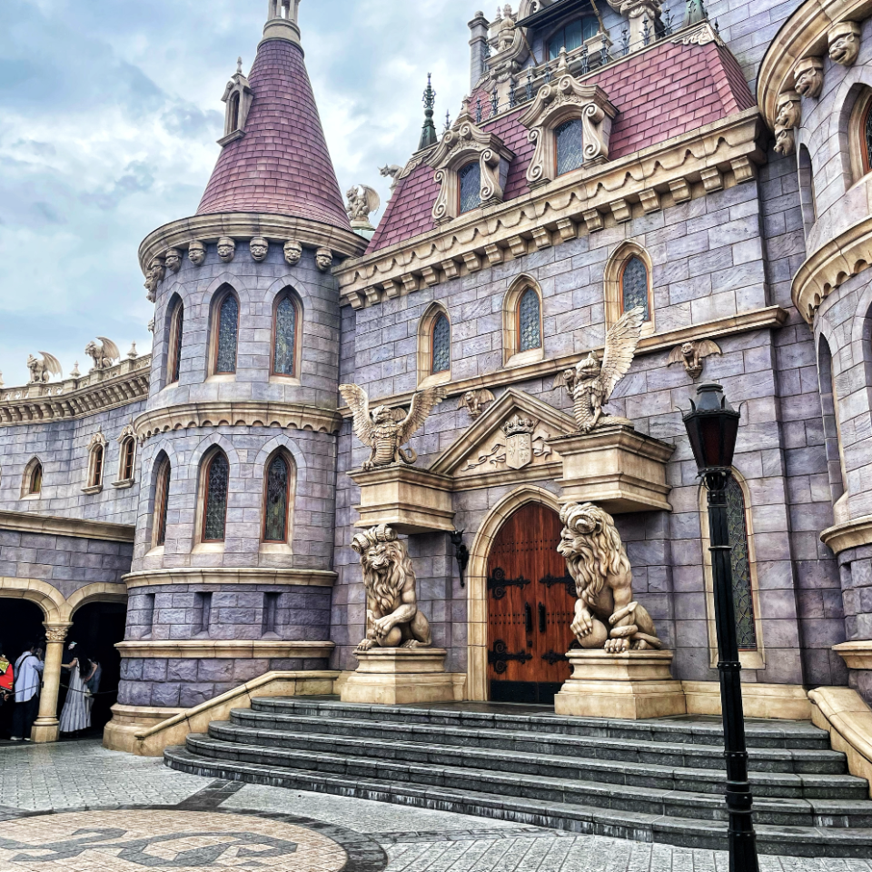 A Gothic-style castle with intricately carved details, featuring two large lion statues flanking the wooden entrance door. The sky is partly cloudy