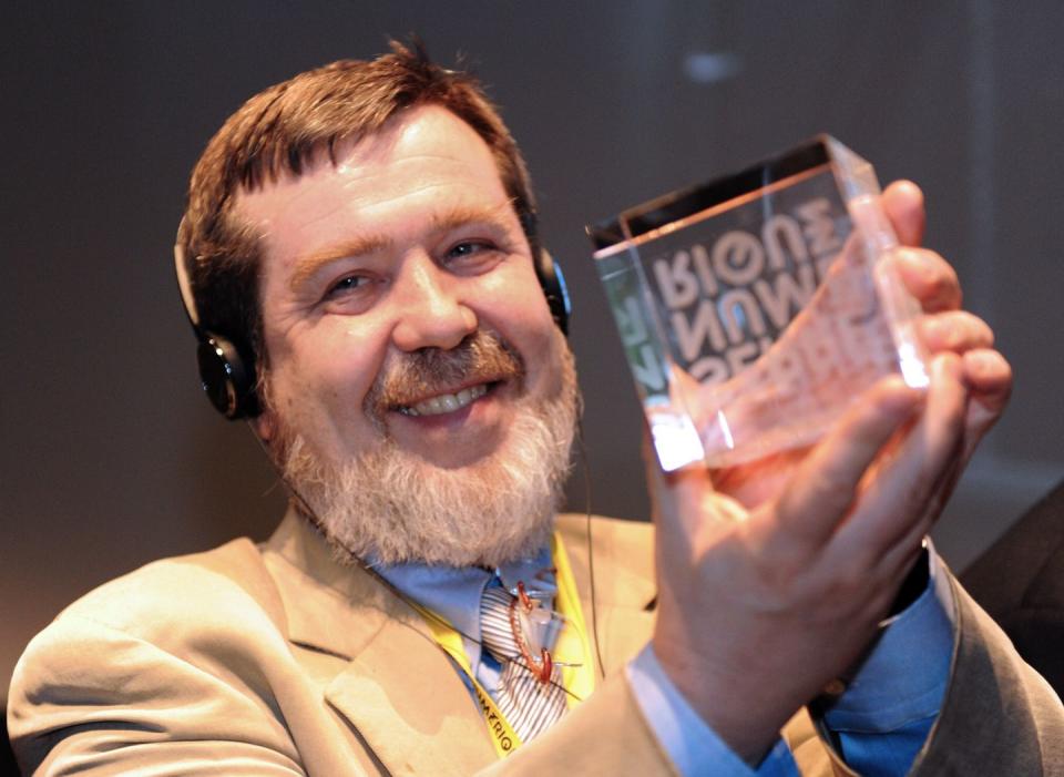 alexey pajitnov, wearing a tan suit and headphones, holds a glass cube trophy and smiles
