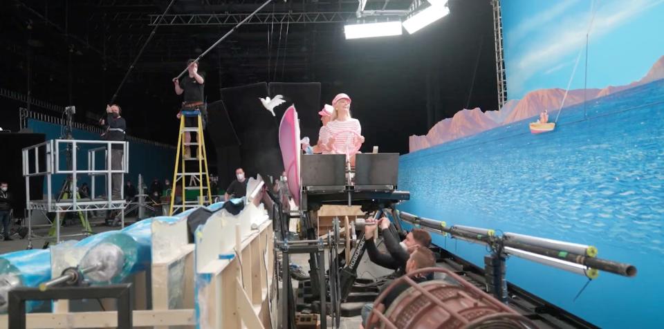 A behind-the-scenes still showing an aquatic-themed set where Margot Robbie and Ryan Gosling are sitting on a boat while set workers film them from the side.