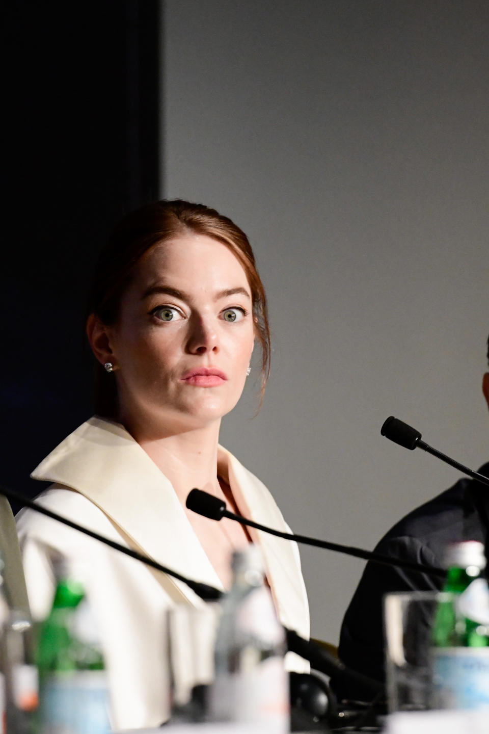 Emma Stone at a press conference, wearing a white blazer, with an attentive expression, seated at a table with microphones and water bottles