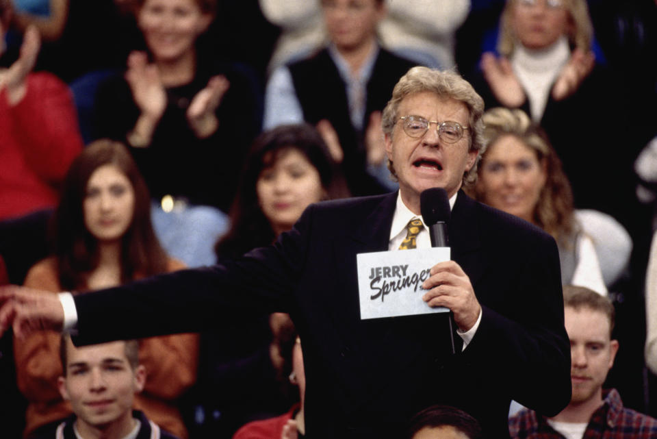 Jerry Springer pointing to something off screen as he talks into the microphone with the audience behind him on "the Jerry Springer Show"