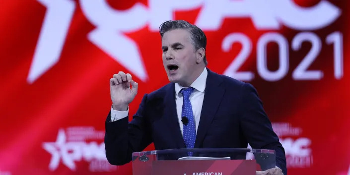 Tom Fitton speaking behind a podium in front of a red background