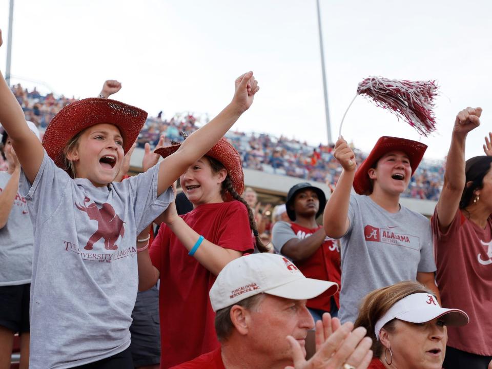Children wearing red cowboy reds and shirts with their team's emblem are chearing in front of a crowd