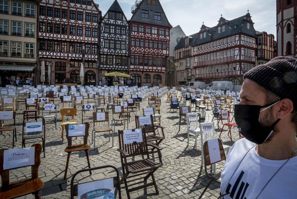 FILE - In this April 24, 2020 file photo, a man with a face mask watches empty chairs with names of bars and restaurants on the Roemerberg square in Frankfurt, Germany. More than 50,000 people have died after contracting COVID-19 in Germany, a number that has risen swiftly over recent weeks as the country has struggled to bring down infection figures. (AP Photo/Michael Probst, File)