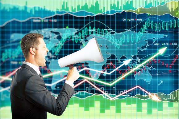 A man in a suit speaks into a megaphone while standing in front of a giant monitor displaying an ascending stock price chart.