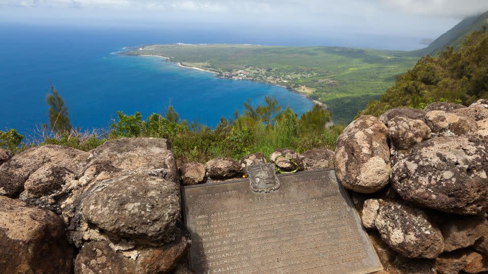 A plaque explains some of the area's history at Kalaupapa Lookout. The peninsula emerges at the base of towering sea cliffs. - Craig Ellenwood/Alamy Stock Photo