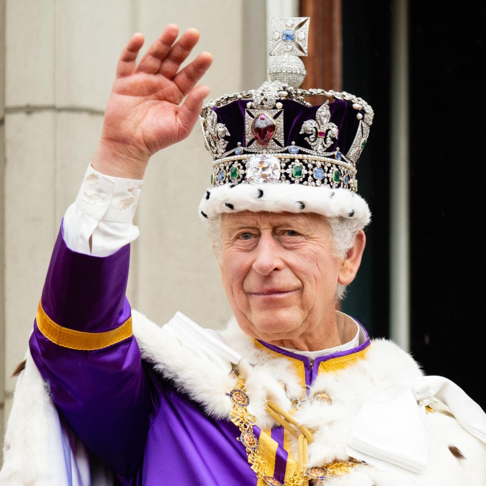 King Charles III, dressed in royal regalia with a jeweled crown and ornate robe, waves during a public appearance