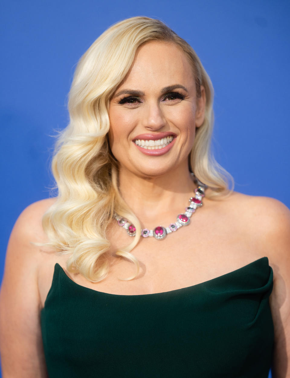 Rebel Wilson at an event wearing a strapless emerald dress and a multicolored gemstone necklace