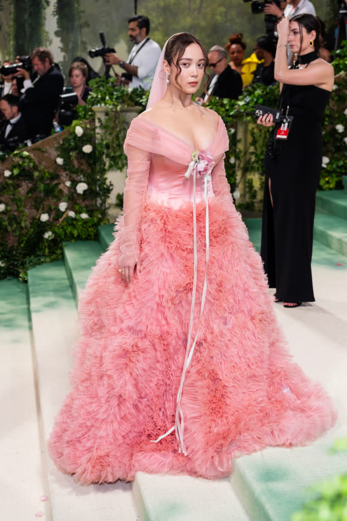 Laufey in a ruffled gown with off-the-shoulder sleeves, standing on a carpeted event. Notable bow detail at waist