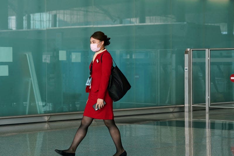 A Cathay Pacific flight attendant wearing a face mask and dressed in a red skirt suit walks through an airport hall.
