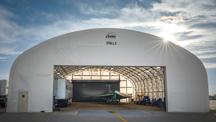     A jet plane with pale green hoods and neat, sharp edges and angles rests under the hangar canopy, culminating in the bright sun over the right side of the hull, against a cloudy blue sky.  