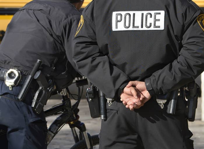 Two officers stand side by side from the neck down, one wears a jacket that says "police" and has their hands folded behind their back; their guns, handcuffs, and utility belts are visible