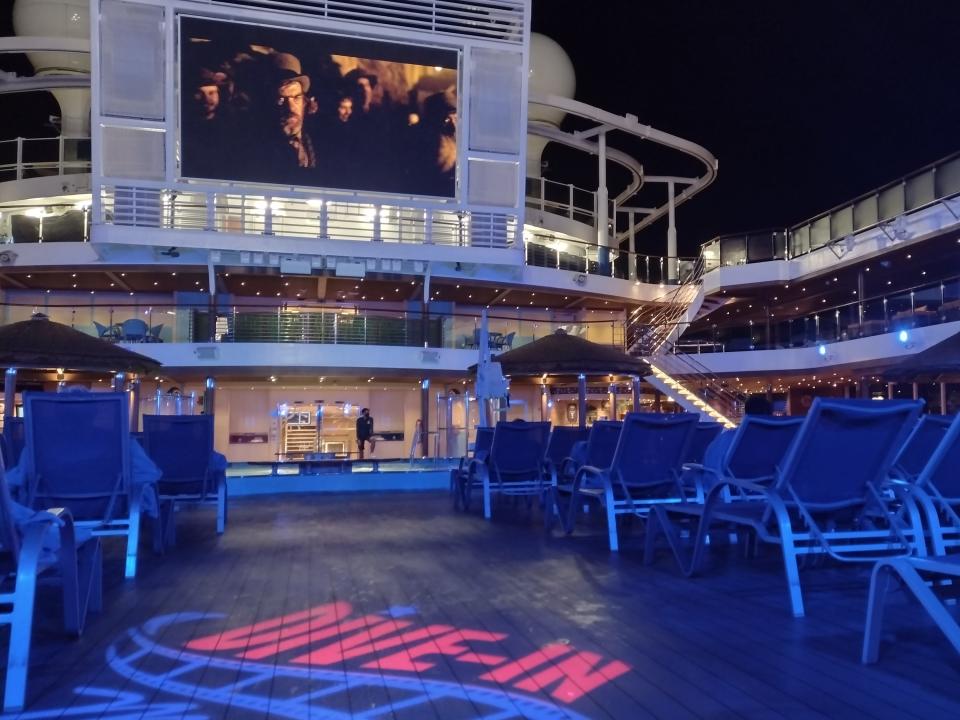 A movie screen on a cruise ship with lines of lounge chairs on the top deck.