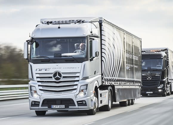 A Mercedes-Benz tractor-trailer truck traveling on a highway. The driver is holding his hands behind his head to show that the vehicle is autonomous.