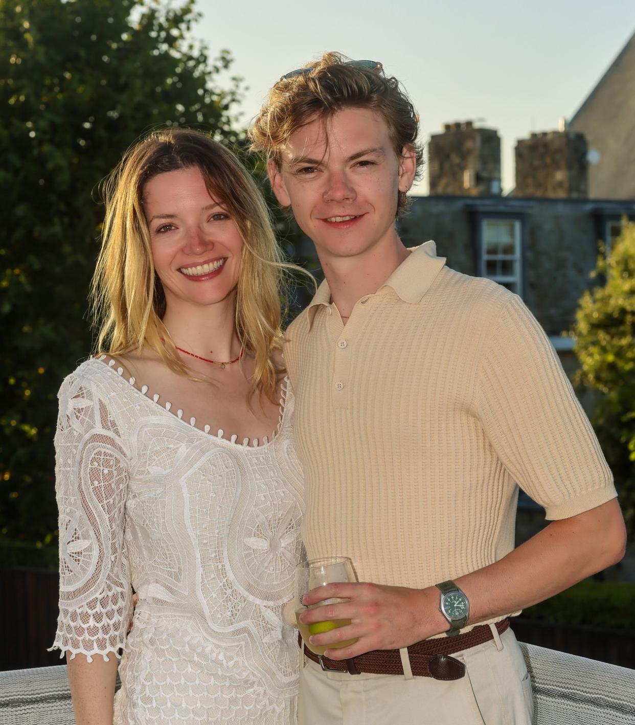 talulah riley and thomas brodie sangster, a young man and woman stand hugging as they smile at the camera, she has blonde hair worn down and wears a white dress, he has light brown curly hair and wears a beige top and trousers