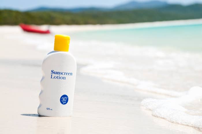 A sunscreen lotion bottle with SPF 30 rests on a sandy beach with gentle waves and a blurred red kayak in the background