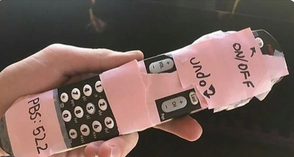 TV remote control with handwritten labels on pink tape for buttons like ON/OFF and PBS: 522, held in someone's hand, suggesting customization for ease of use