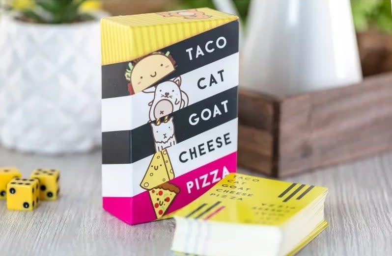 A card game named "TACO CAT GOAT CHEESE PIZZA" with whimsical illustrations, set on a table next to dice