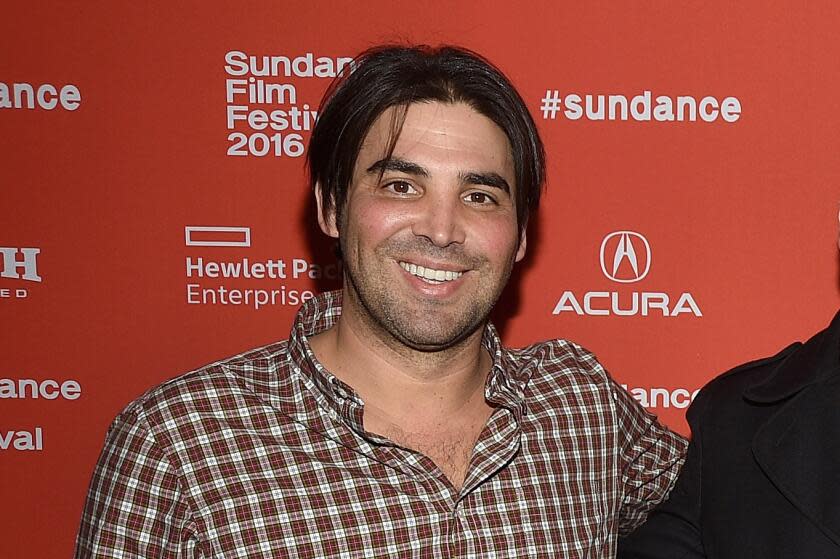 A man with dark medium-length hair wearing a checkered shirt and smiling against a red backdrop