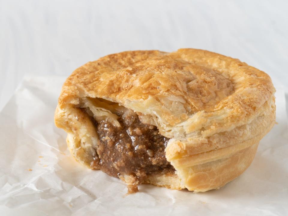 A white napkin holding a small pastry with a meat filling spilling out of it on a white table