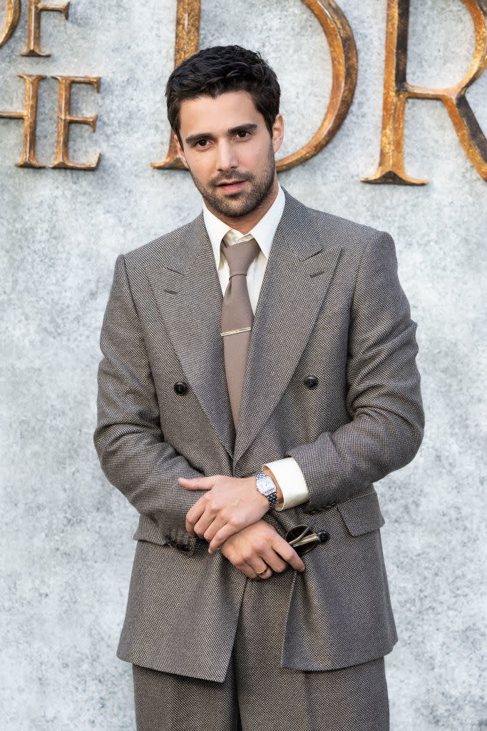 Simone Ashley in a double-breasted suit with a tie, standing in front of a textured background