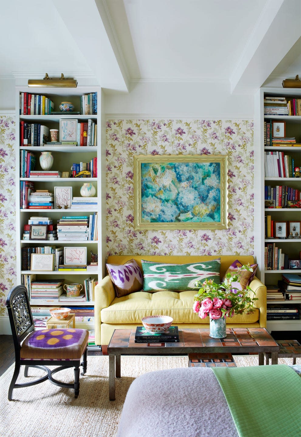 sitting area with flowered wallpaper, a light yellow sofa with ikat accent pillows and flower artwork above it, tall bookshelves flank sofa, cocktail table with a vase of flowers, chair with purple cushion, natural rug