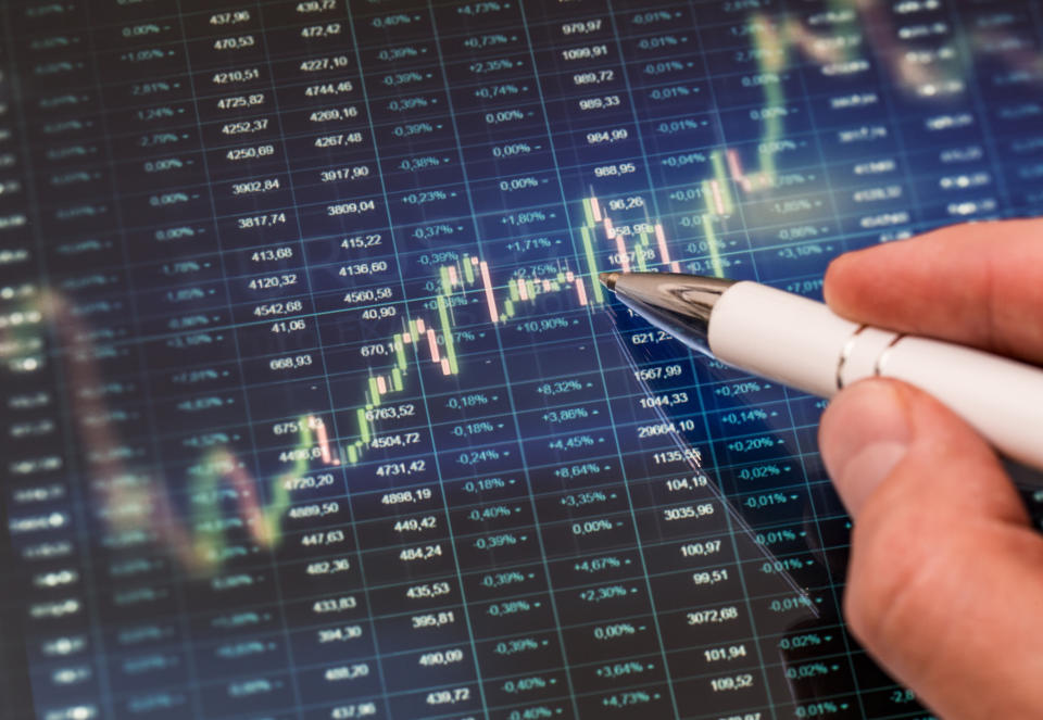 A pen tracing along a candle stick chart with stock tickers in the background