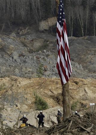 A flag flies at half staff as workers look out over the mud and debris as search work continues from a massive mudslide that struck Oso near Darrington, Washington April 2, 2014. REUTERS/Jason Redmond
