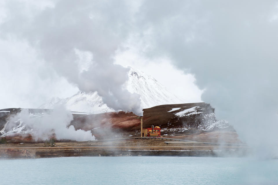 Steam billows from a geothermal plant painted red, with a hill in the background partially covered in snow.