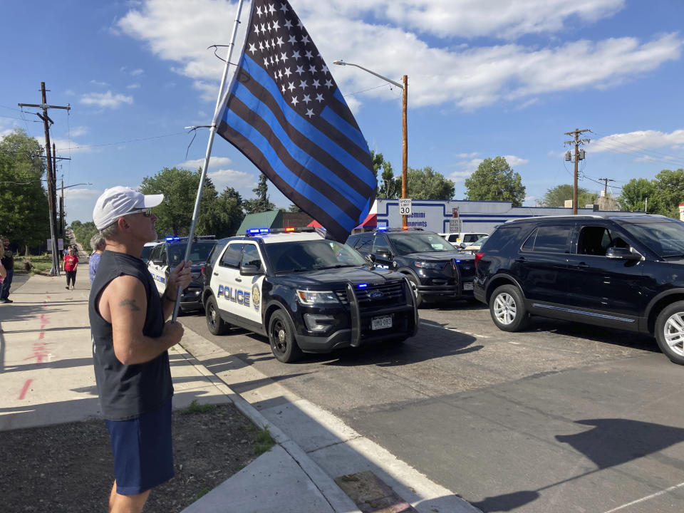 John Garrod, of Arvada, stands holding a blue line flag at the beginning of a line of about 30 police cars lined up for a procession in honor of the officer who was fatally shot in Arvada, Colo., on Monday, June 21, 2021. A gunfight between two men and police officers at a shopping district in a Denver suburb left an officer and one of the suspects dead, authorities said Monday. (AP Photo/Colleen Slevin)