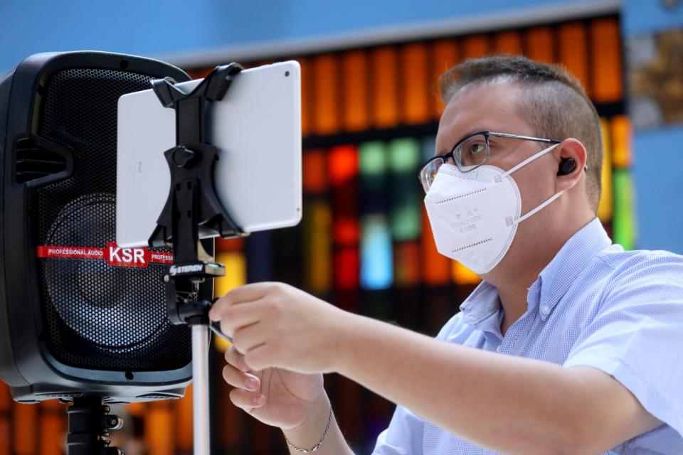 A man in a mask looks at a tablet computer on a tripod stand next to a speaker