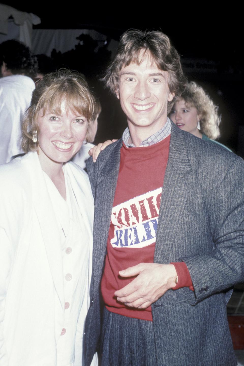 Martin Short and Nancy Dolman smiling while posing for a photo. Martin wears a casual suit jacket over a graphic tee, and Nancy is in a light-colored outfit