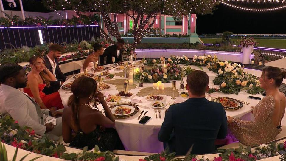 A group shot of Love Island contestants sitting around a round dinner table, with food on plates, flowers and a white tablecloth.