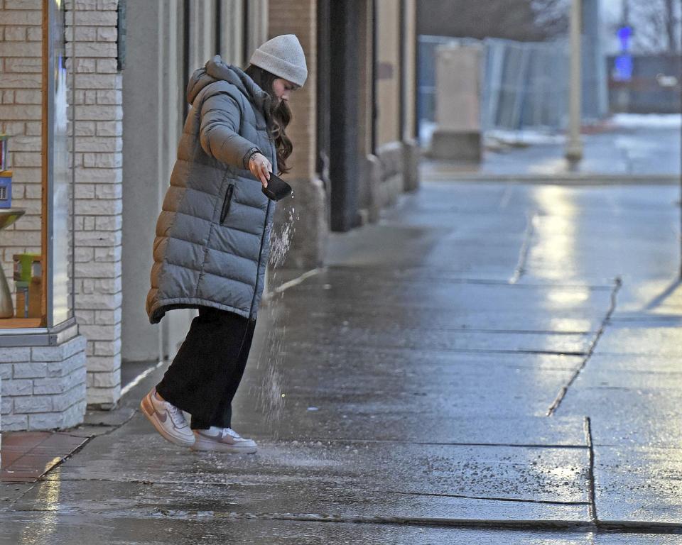 Mariah Sanders sprinkles rock salt to melt the ice covered sidewalk outside the Hi Honey salon downtown Bismarck, N.D., on Tuesday morning, Dec. 26, 2023, as ice covered streets and sidewalks made walking and driving difficult. Sanders said she was expecting a customer, but others had called in to postpone due to weather conditions. (Tom Stromme/The Bismarck Tribune via AP)