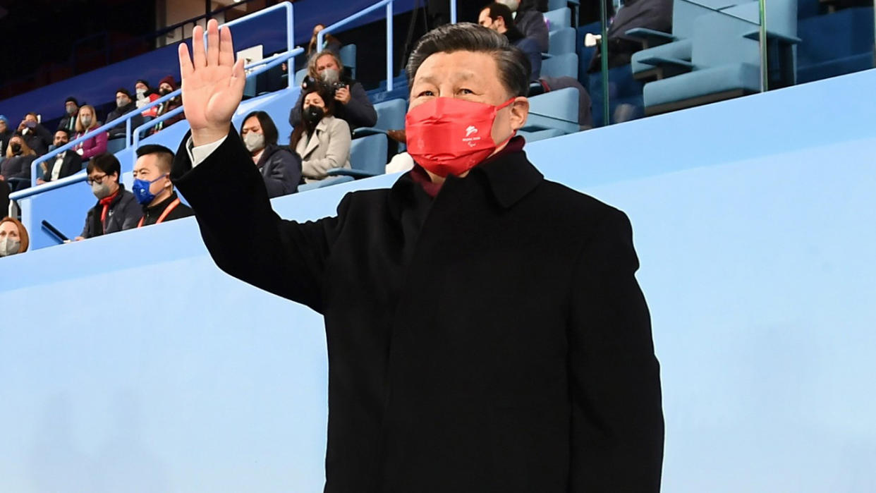 Chinese President Xi Jinping, wearing a black coat and red face mask, stands and waves in a stadium with a few dozen spectators visible seated behind him.